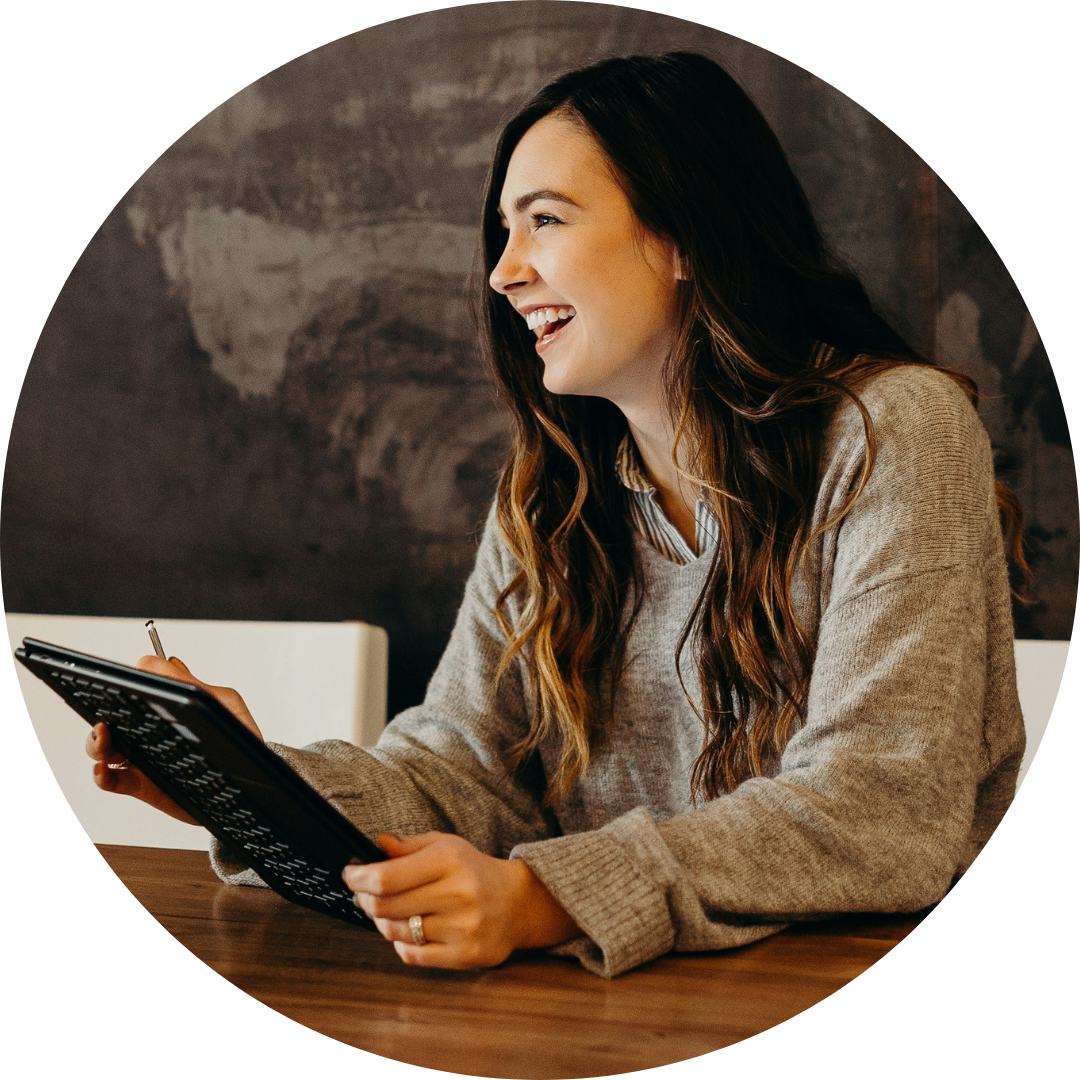 A woman with long hair smiles and laughs while sitting at a table, holding a pen and tablet. She is wearing a gray sweater in a casual setting with a dark-toned background. As an entrepreneur possibly starting a business, she appears to be engaged and enjoying the moment.