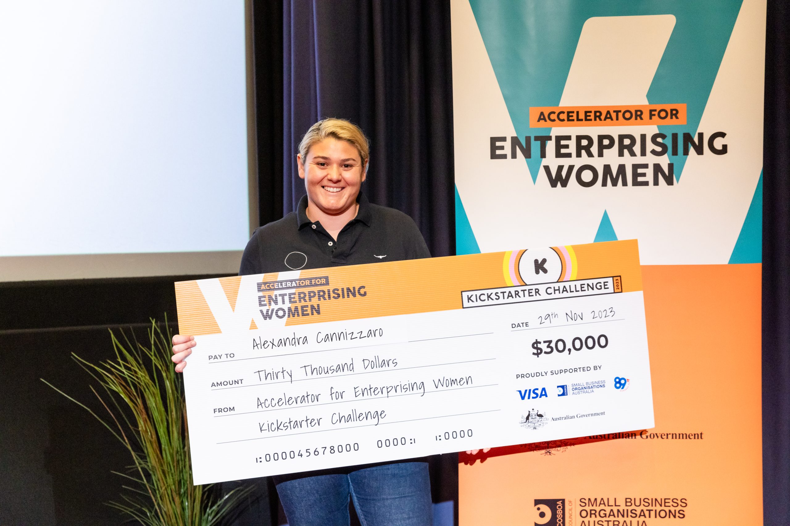 A person stands smiling, holding a large check for thirty thousand dollars. The check is addressed to "Alexandra Cannizzo" from the "Accelerator for Enterprising Women Kickstarter Challenge." The background features a banner with the same text and event branding, celebrating women in business.