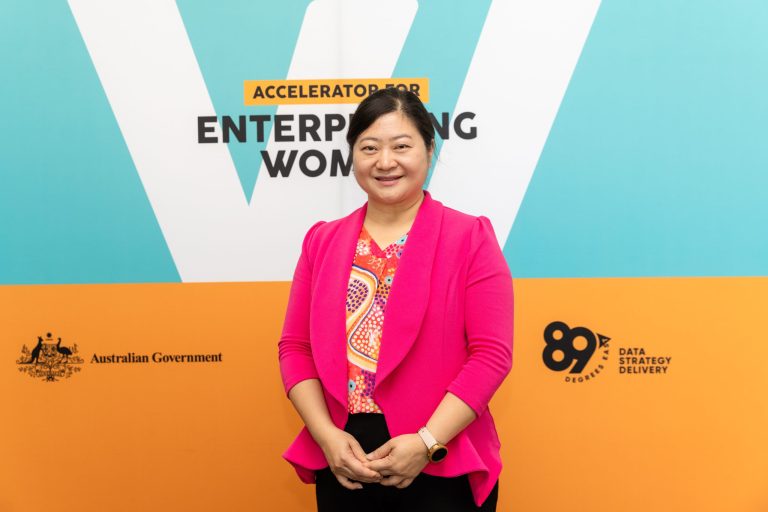 A woman wearing a vibrant pink blazer and a multicolored blouse stands smiling against a backdrop that reads "Accelerator for Enterprising Women." Australian Government and 89 Degrees East logos are visible on the background, promoting business ideas for women entrepreneurs starting a business.