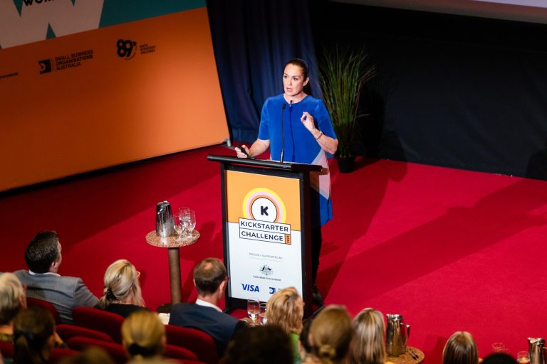 A person stands at a podium delivering a speech on a red-carpeted stage. The podium features signs for the Kickstarter Challenge, Small Business Festival, and various sponsor logos. The audience listens attentively to inspiring business ideas for women. A large orange and blue backdrop is behind them.