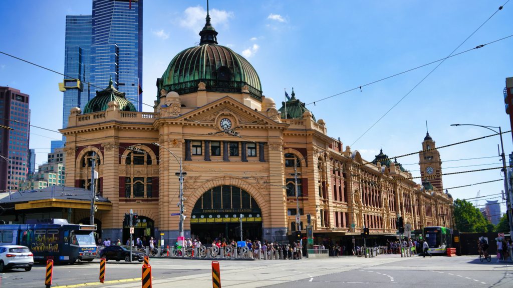 A busy street scene featuring the historic Flinders Street Station in Melbourne, Australia. The station's iconic yellow facade and green domes are prominent, with trams and pedestrians visible in the foreground. Modern skyscrapers in the background set a dynamic stage for entrepreneurs starting a business.