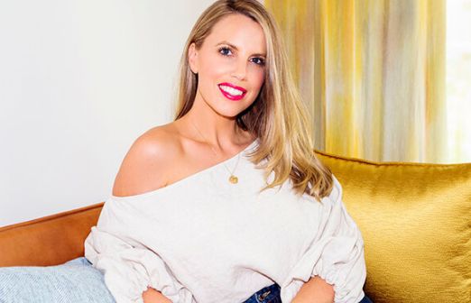 A woman with long blonde hair and bright red lipstick is sitting on a sofa. She is wearing an off-the-shoulder white blouse and dark jeans. Behind her, there is a yellow cushion and a curtain with yellow and white stripes, setting the perfect backdrop for discussing business ideas for women entrepreneurs.