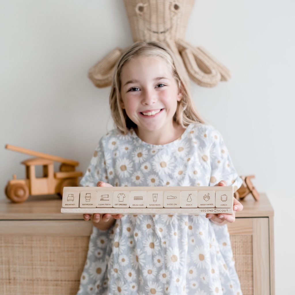 A smiling young girl with blonde hair holds a rectangular box with various icons and text, perhaps sparking early business ideas for women. She wears a gray dress with white and yellow daisy patterns. Wooden toys are displayed on a cabinet behind her, with a cheerful woven wall decoration above.