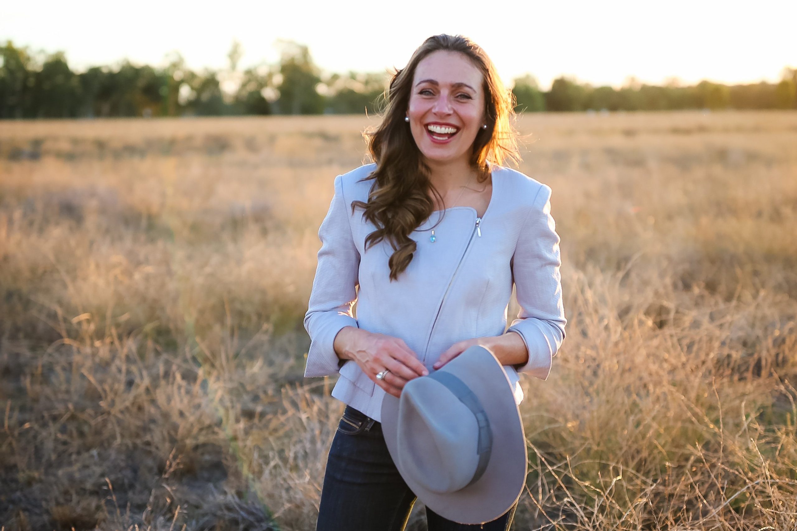 A woman with long wavy hair smiles brightly while standing in a sunlit field of tall, dry grass. An inspiring symbol for women in business, she is wearing a light jacket and dark pants, holding a light gray hat in front of her. The clear sky and sunset horizon serve as a backdrop to her entrepreneurial spirit.