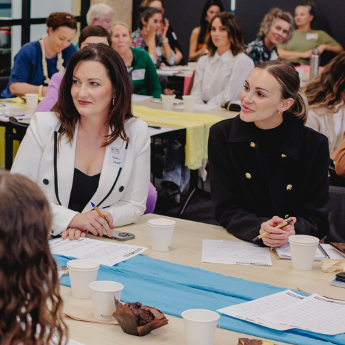 A group of people sitting at tables in a bright room, participating in a workshop or seminar on business ideas for women. Two women in the foreground are listening attentively, one in a white blazer and the other in a black jacket. Various papers, pens, and cups are on the tables.