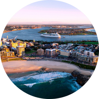 Aerial view of a coastal city featuring sandy beaches, waves, and urban buildings. A cruise ship navigates the harbor under a clear sky at sunset, with the cityscape and ocean extending into the distance.
