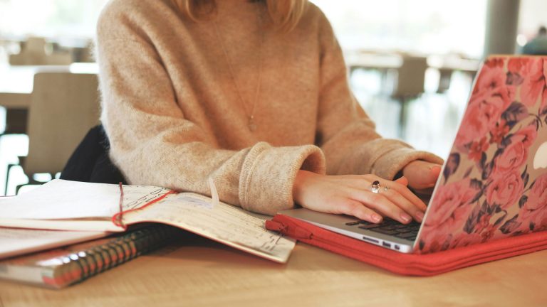 A person wearing a beige sweater types on a laptop with a pink floral design, possibly researching business ideas for women. An open notebook with handwritten notes and a spiral-bound book are on the table next to the laptop. The background features basic, out-of-focus furniture.
