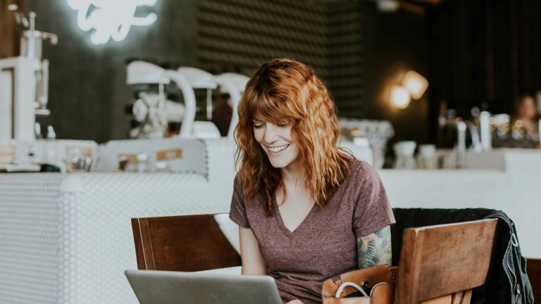 A woman with wavy red hair and a purple shirt is sitting in a cozy cafe, smiling at her laptop screen. An entrepreneur starting a business, she finds inspiration in the vibrant background that features a coffee bar with various equipment and a neon sign.