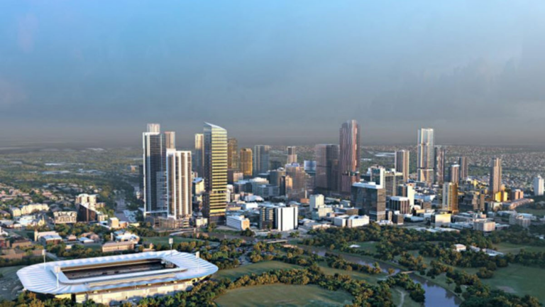 Aerial view of a modern cityscape with numerous skyscrapers and tall buildings of varying designs and heights. The city is bordered by green parks and a river. In the foreground, there is a large sports stadium surrounded by lush greenery, exemplifying an inspiring hub for women in business.