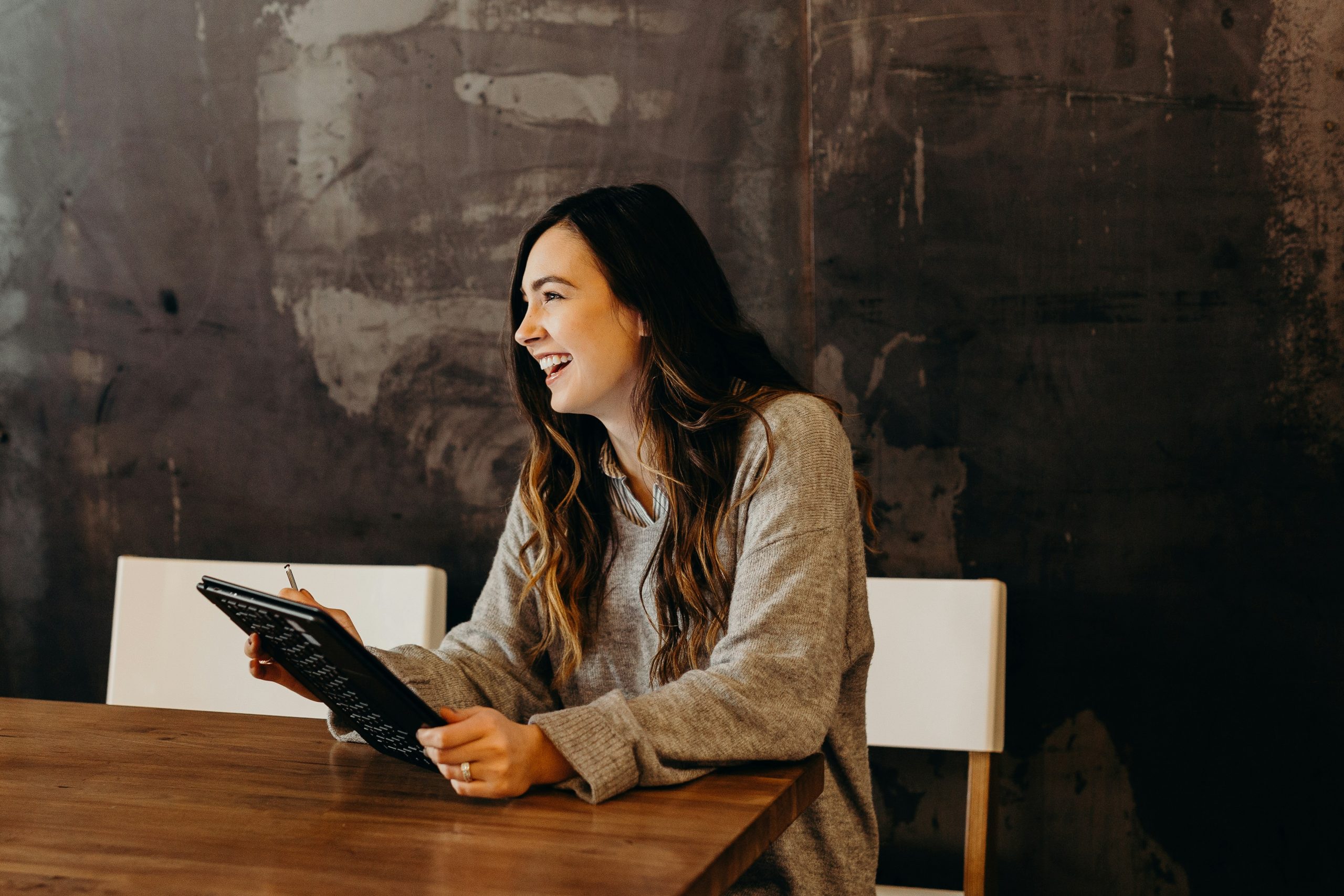 A woman with long brown hair sits at a wooden table, smiling and holding a black tablet in her hand. She is wearing a grey sweater, and the background features a dark, textured wall. The atmosphere appears to be relaxed and cheerful—perfect for discussing women in business.