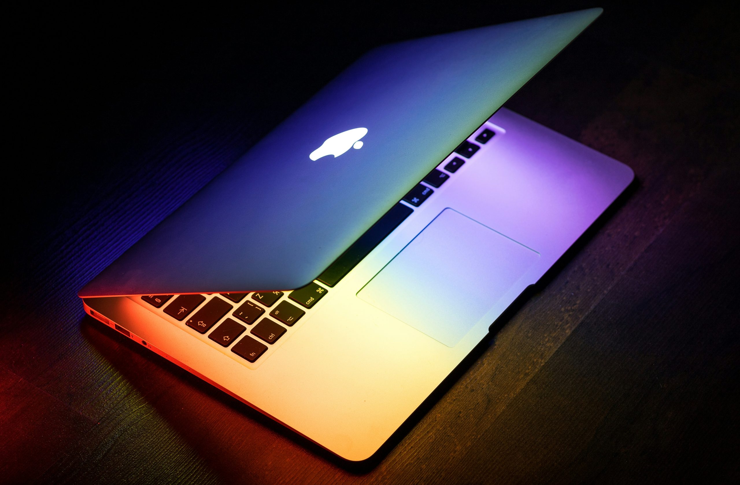 A partially open laptop with a glowing apple logo, emitting a spectrum of colorful light across its keyboard and surface, sits on a dark desk. The vibrant display inspires innovative business ideas for women, highlighting the potential for female entrepreneurs starting a business.