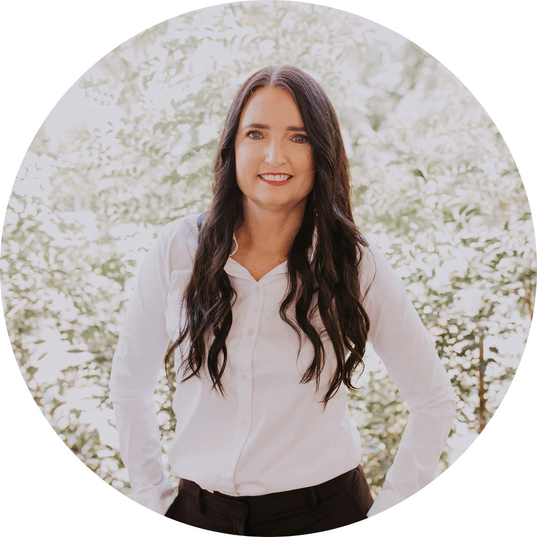 A woman with long, dark hair is smiling and wearing a white blouse and dark pants. She is standing in front of a backdrop of lush green foliage.