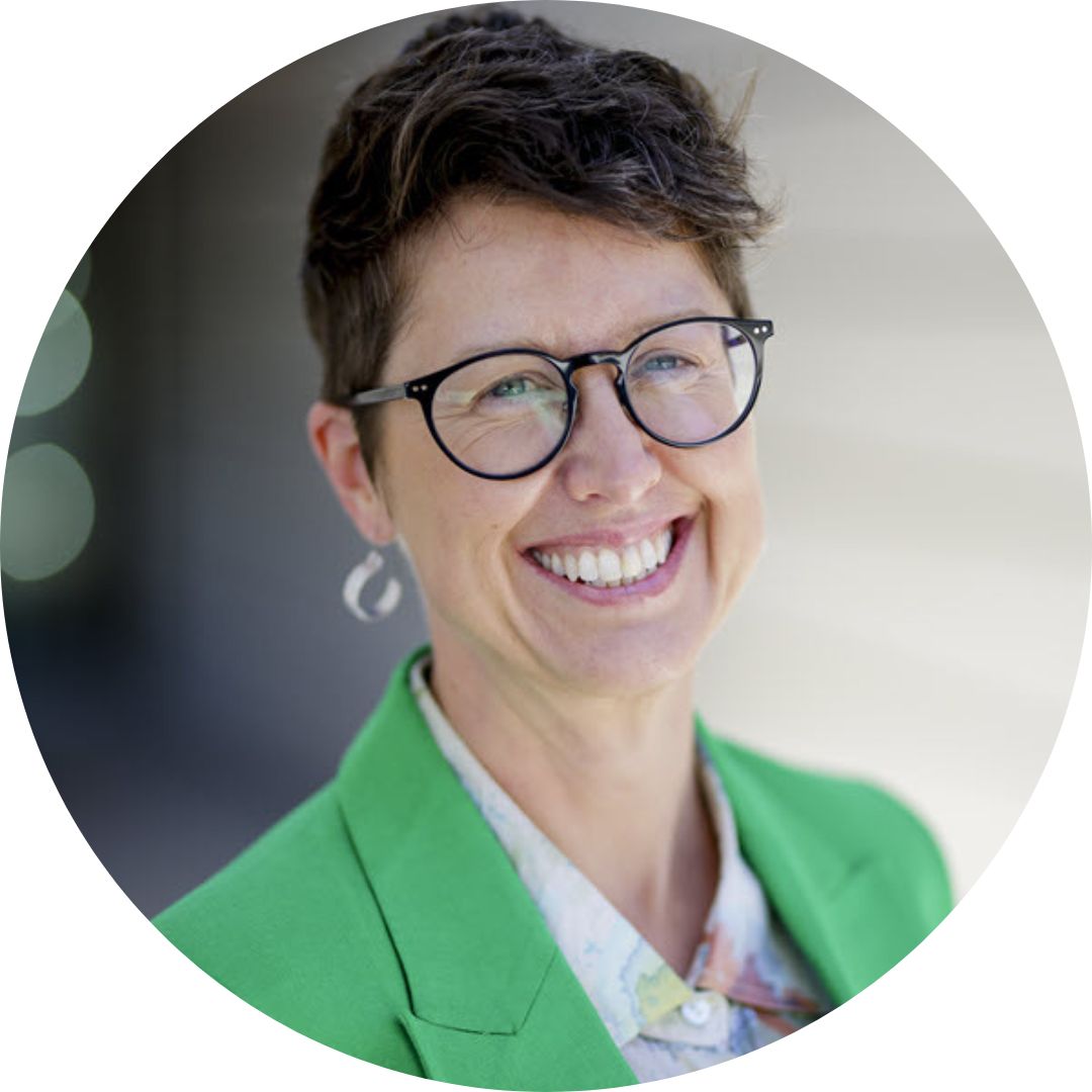 A person with short hair, black glasses, and a beaming smile is dressed in a bright green blazer over a light-colored shirt. The background is blurred, focusing attention on the person's joyful expression.