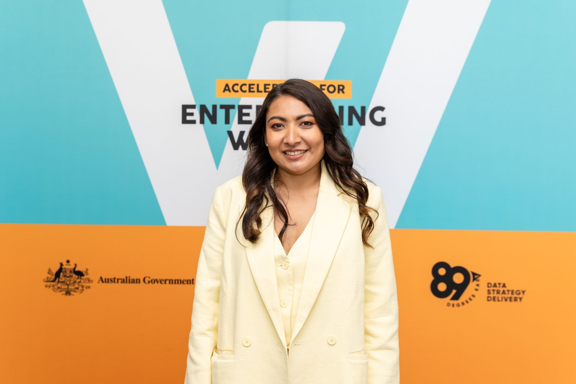 A woman with long dark hair, wearing a light yellow blazer, smiles while standing in front of a backdrop with bold colors and text that reads "Accelerator for Enterprising Women," along with logos for the Australian Government and 89 Degrees East Data Strategy Delivery.