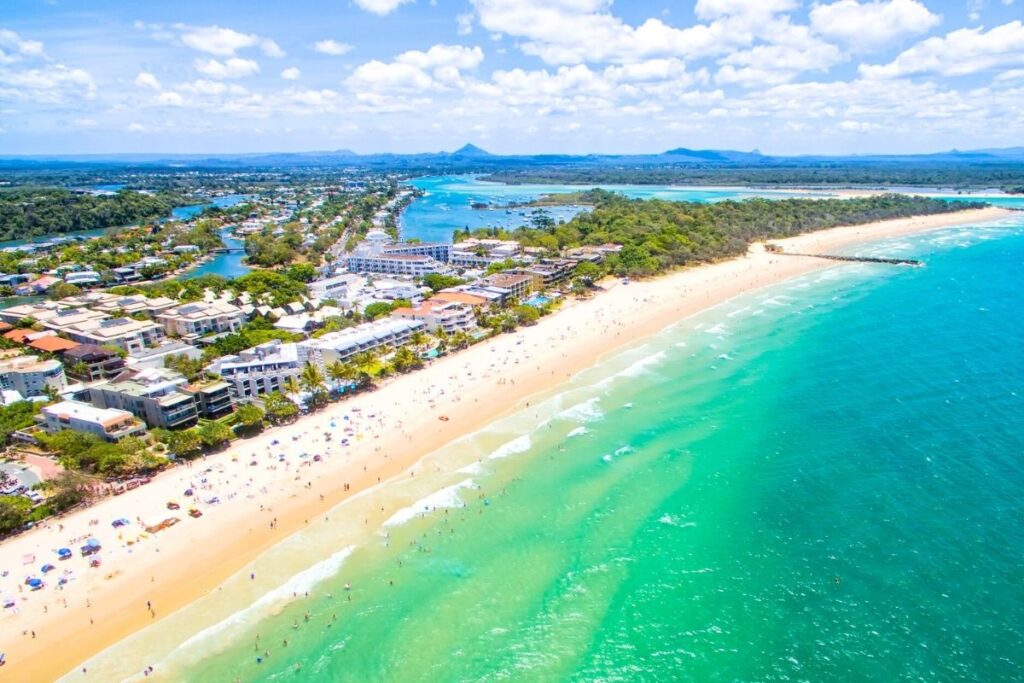 Aerial view of a bustling beach along a turquoise ocean. The sandy shoreline is crowded with sunbathers under umbrellas, while waves gently lap the shore. Buildings and greenery extend along the coast, with a river and mountains visible in the background.
