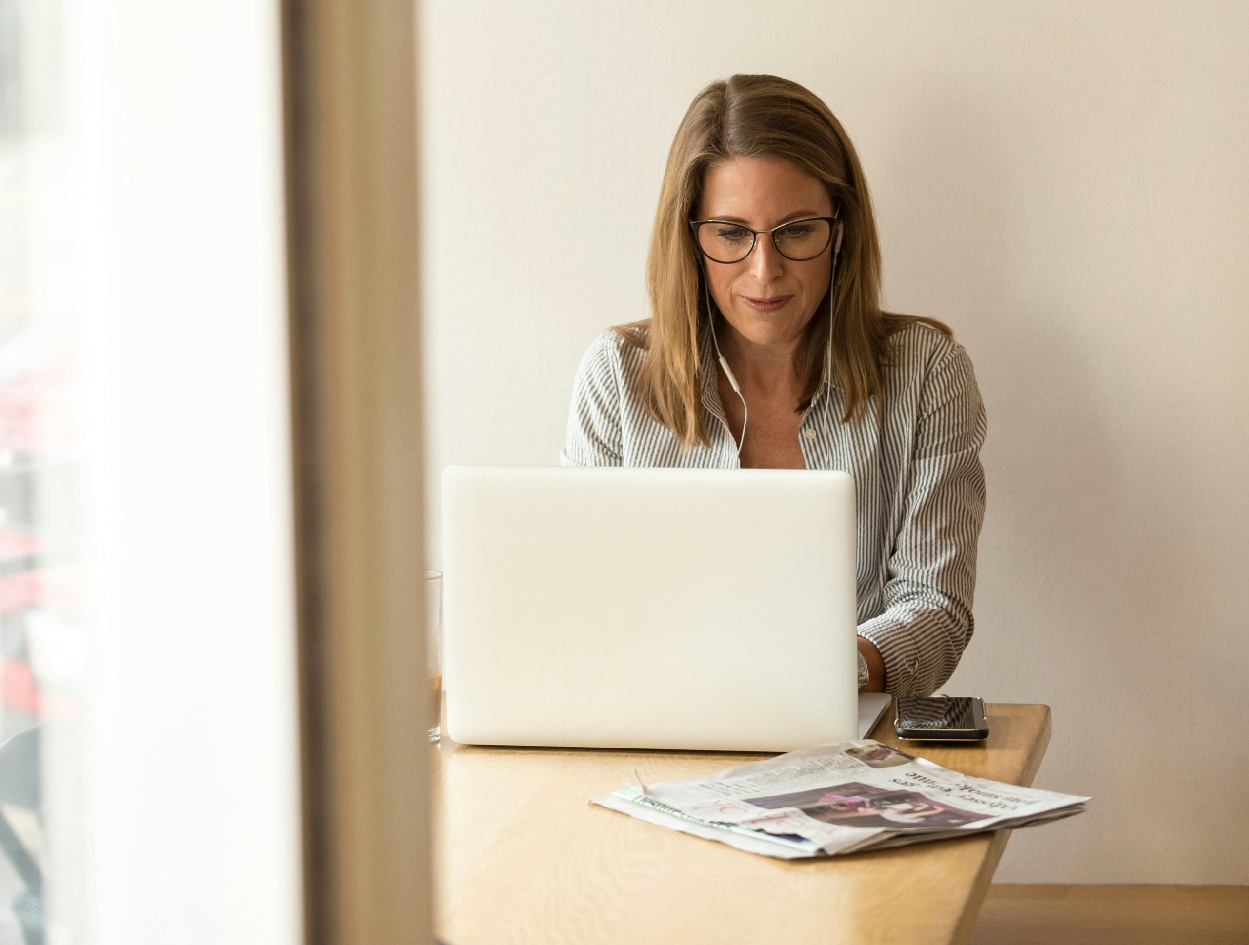 A woman with shoulder-length hair and glasses is sitting at a wooden table, working on a laptop. Newspapers are spread out on the table beside her, and a smartphone is also visible. There's natural light coming through a window on the left side.