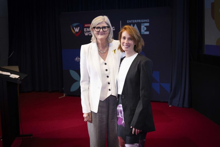 Two women standing on a red carpeted stage, smiling at the camera. One wears a white blazer and patterned pants, the other a black blazer and white skirt. Behind them is a dark blue backdrop with logos and text related to an event.