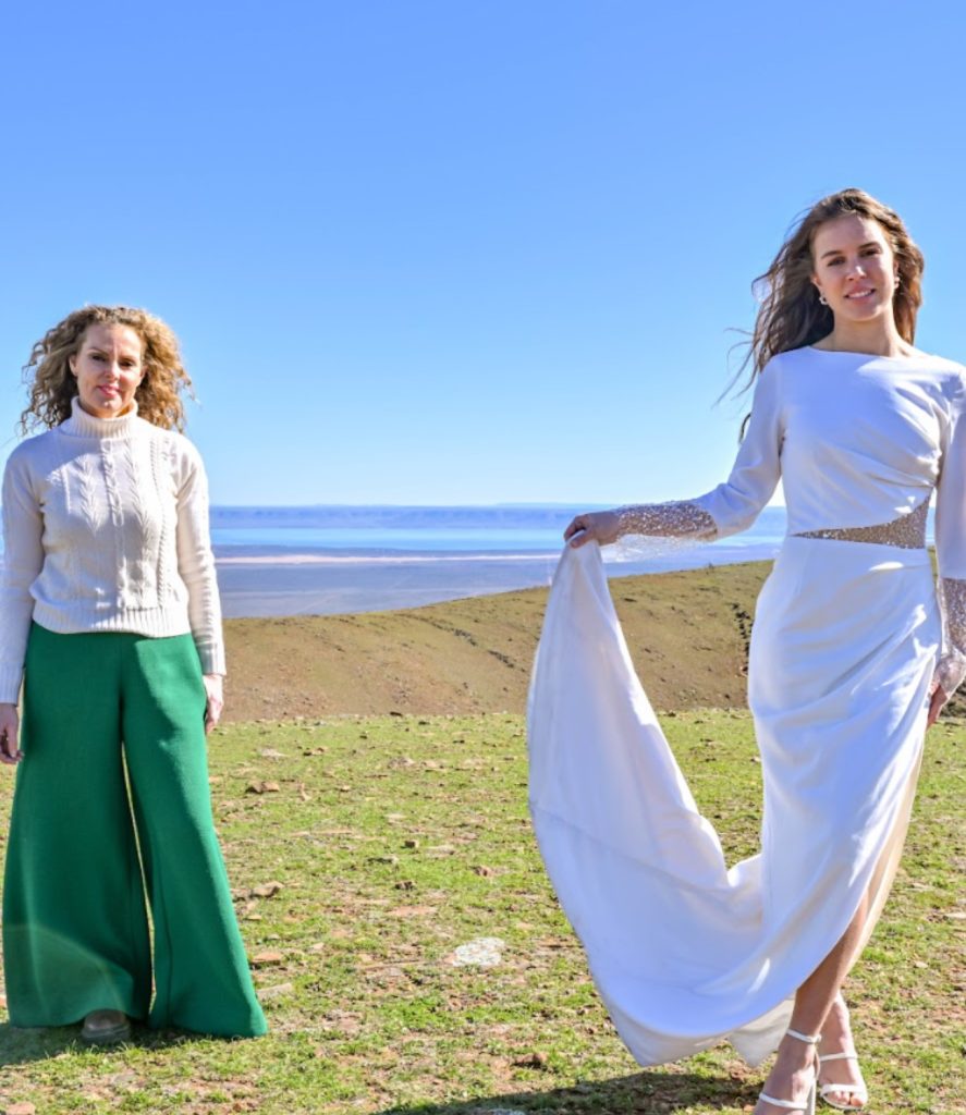 Two women stand outdoors in a grassy area. One wears a long-sleeved white dress with lace accents, holding the skirt. The other wears a white sweater and green pants. A vast view of water and clear blue sky is in the background.