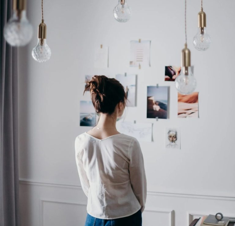 A person with their hair in a bun, wearing a light long-sleeve shirt, stands facing a wall covered with various photographs and notes. Several hanging light bulbs are visible above, and a small section of a table is in the lower right corner.