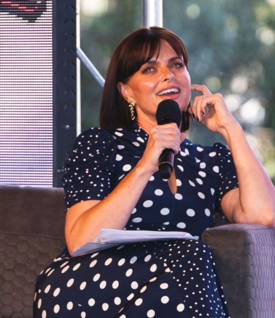 A woman in a navy blue polka dot dress holds a microphone while sitting on a stage. She is smiling and appears engaged. There's a blurred background with greenery and part of an electronic screen on the left.