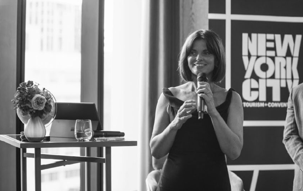 A woman is speaking into a microphone at an indoor event. She stands near a table with a laptop and a floral arrangement. A sign behind her reads "New York City Tourism & Conventions." The photo is in black and white.