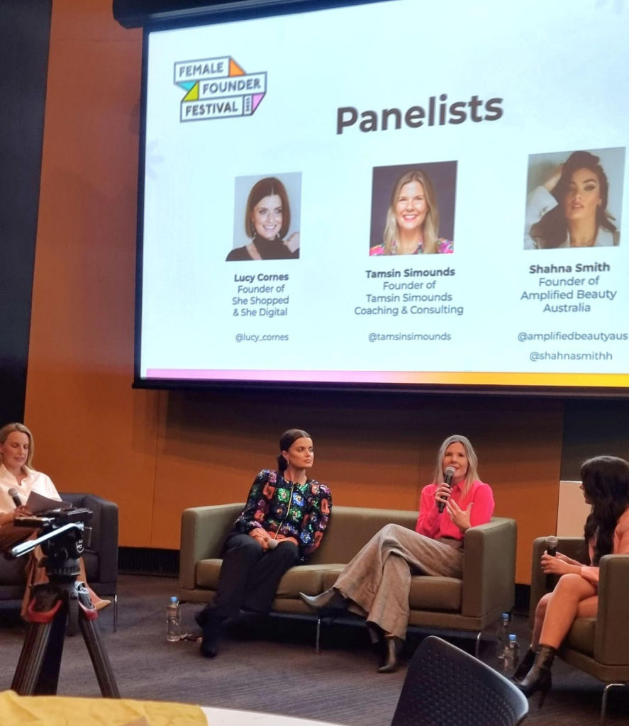 Five women sit in a panel discussion at the Female Founder Festival. A large screen behind them displays photos and names of three panelists: Lucy Cornes, Tamsin Simounds, and Shahna Smith. The seated women hold microphones and engage with the audience.