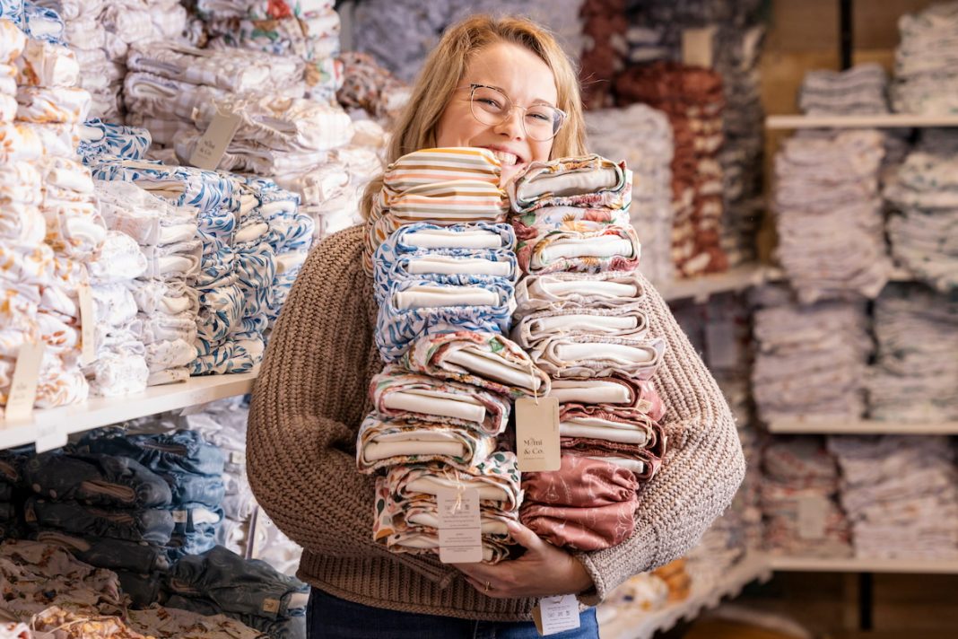 A person wearing glasses and a sweater is smiling while holding a large stack of folded fabrics in a store. Shelves filled with more colorful folded fabrics are in the background.