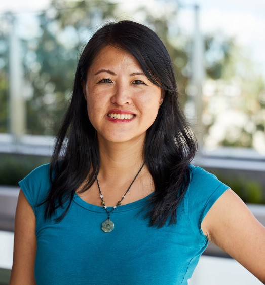 A person with long dark hair is smiling and wearing a blue top and necklace. They are standing outdoors with a blurred background of trees and a railing.