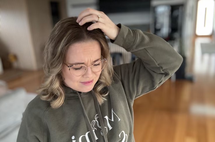 A woman with glasses, wearing a green hoodie, stands indoors with a slightly pained expression. She's touching her head as if in discomfort. The room has wooden floors and blurred background elements, suggesting a home environment.