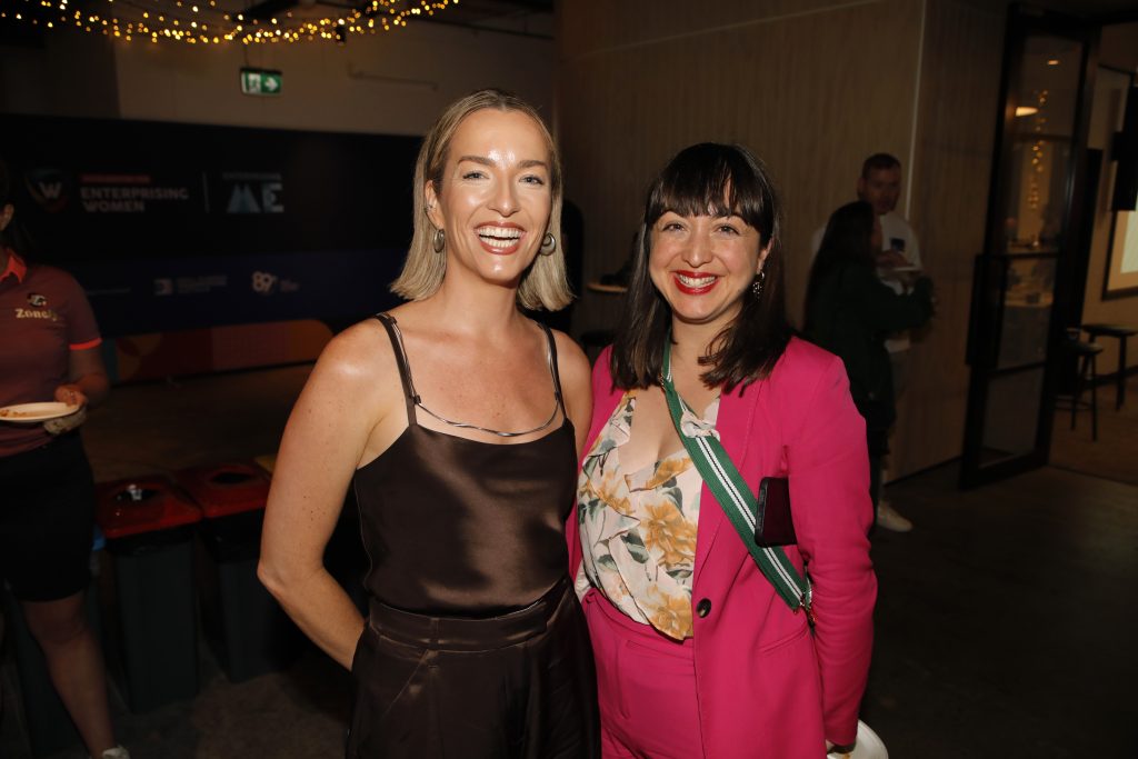 Two women are smiling and standing together at an indoor event. One wears a dark sleeveless top, and the other is in a pink blazer with a floral top. The background has dim lighting and event banners.