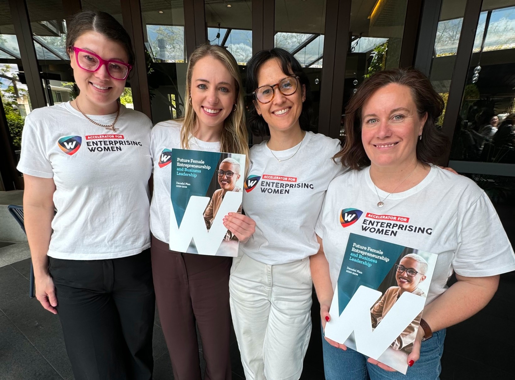 Four women stand together smiling, wearing white "Enterprising Women" T-shirts. Three of them hold magazines featuring the same cover image of a woman and the word "Enterprising Women." Trees and building windows are in the background.