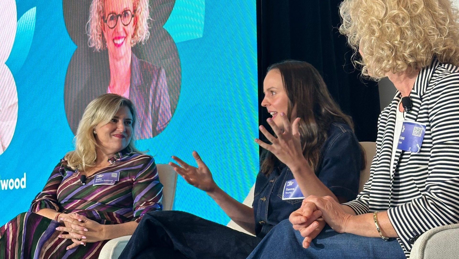 Three people are seated in chairs on stage, engaged in conversation. The background has a large screen with a headshot of one person. They're dressed casually, and one is gesturing animatedly. A small table holds a glass of water.