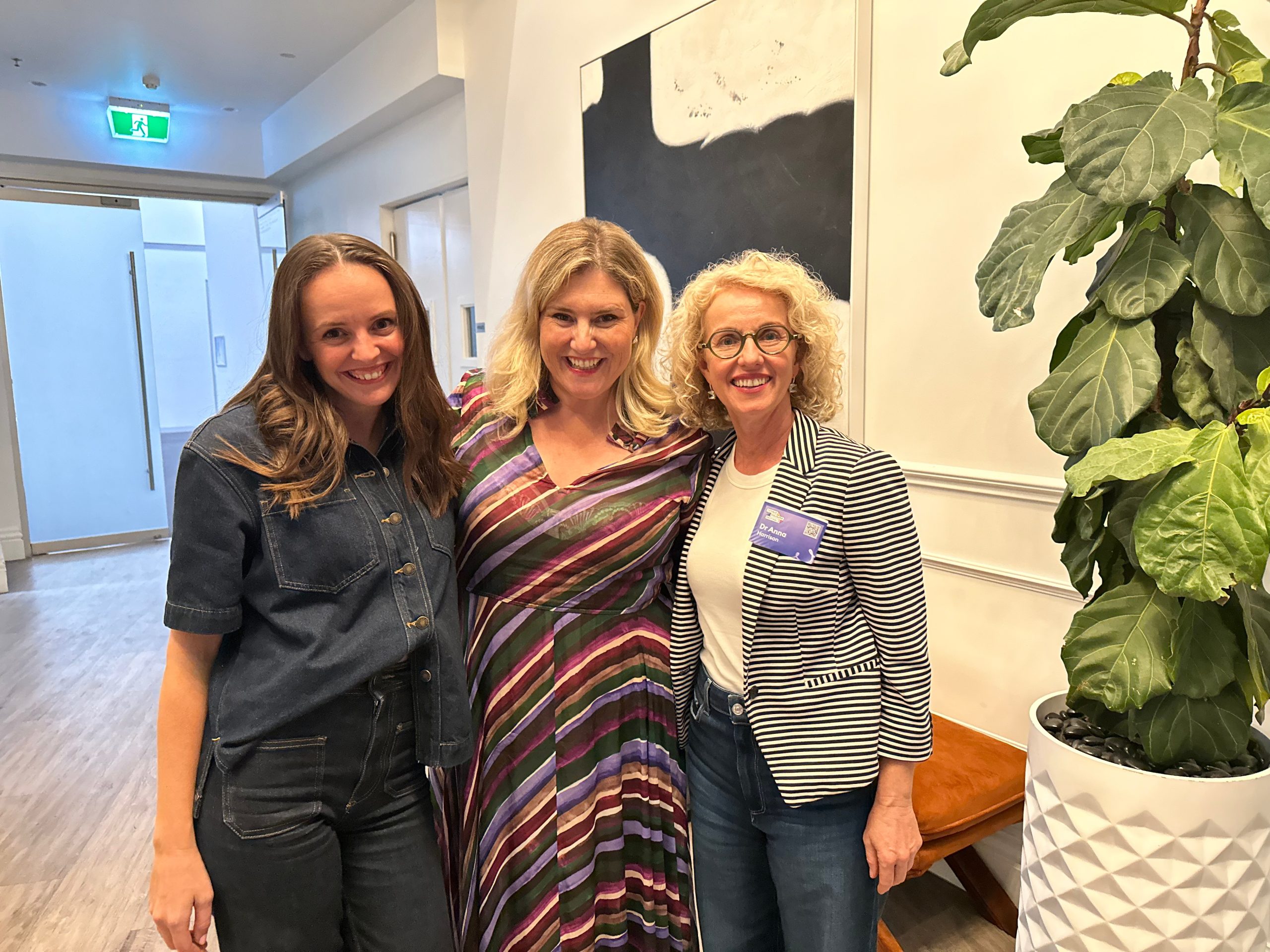 Three smiling women stand together indoors. The first wears a denim outfit, the second a striped dress, and the third a striped blazer with a name badge. They stand beside a large potted plant. A hallway is visible in the background.