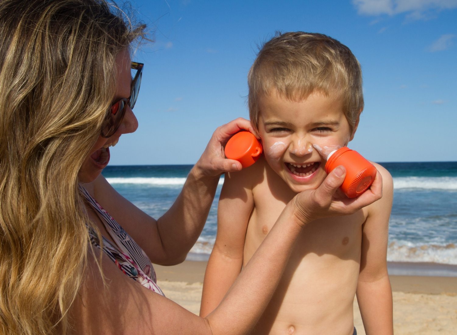 A woman, possibly brainstorming business ideas for women, playfully applies sunscreen to a smiling young boy's face at the beach using bright orange sunscreen applicators. The ocean and sky with a few clouds are visible in the background. The child is shirtless, and both are enjoying a sunny day.