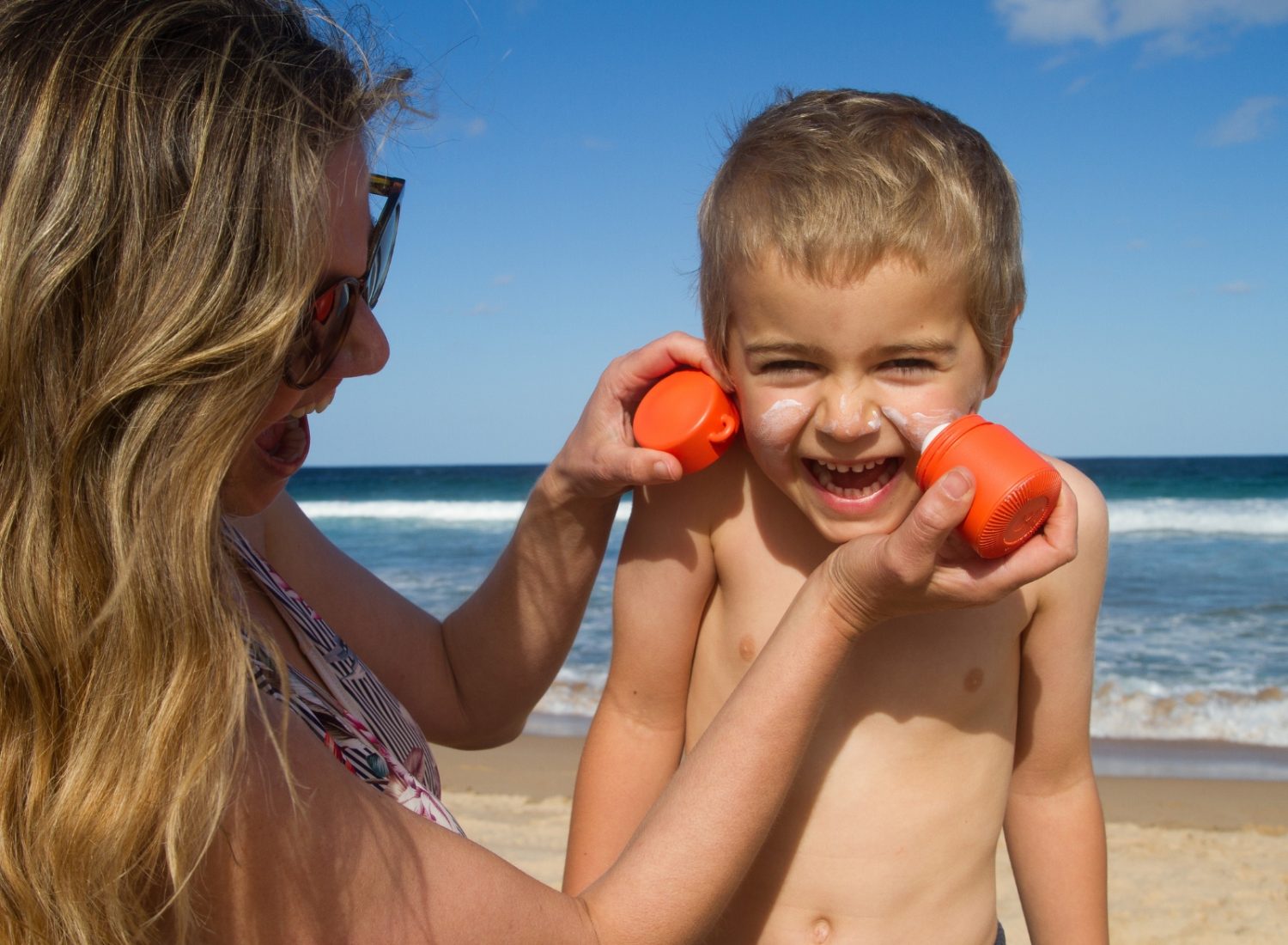 A woman, possibly brainstorming business ideas for women, playfully applies sunscreen to a smiling young boy's face at the beach using bright orange sunscreen applicators. The ocean and sky with a few clouds are visible in the background. The child is shirtless, and both are enjoying a sunny day.